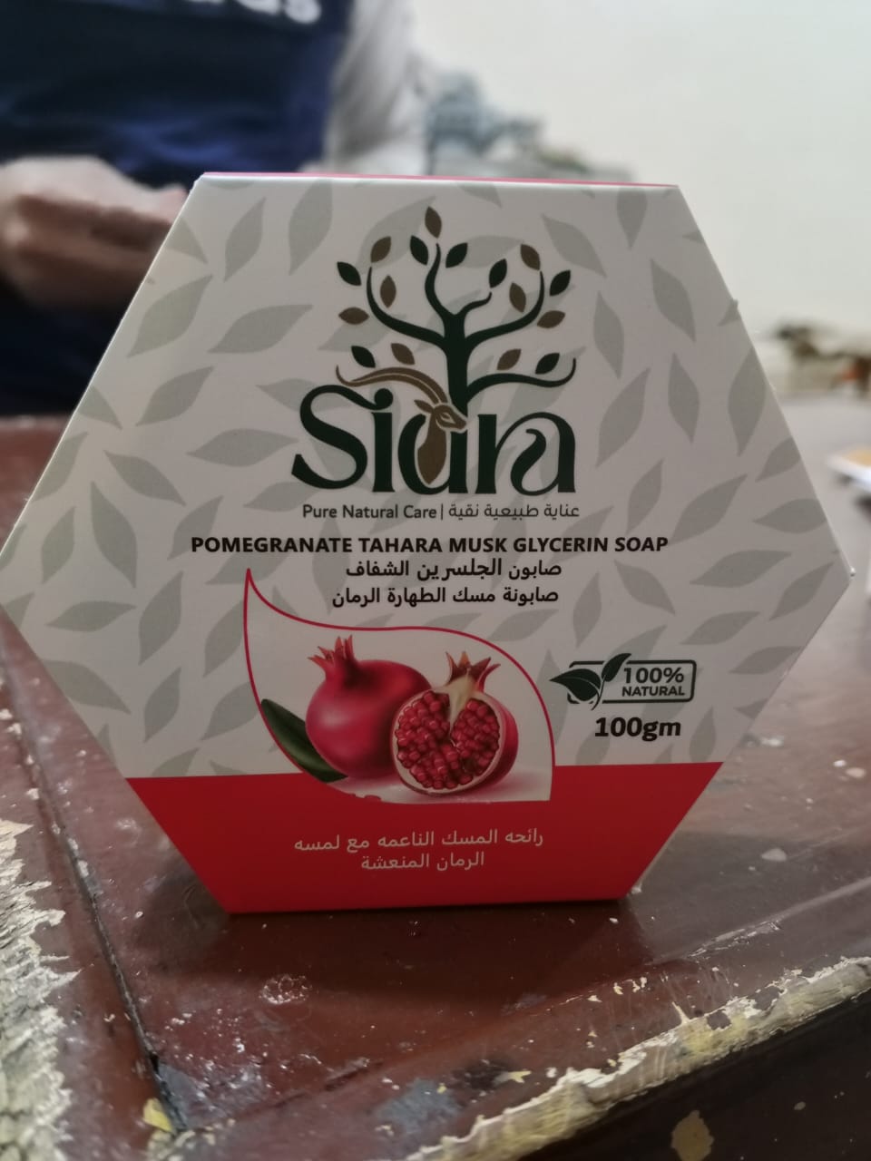 علبة صابونة مسك الطهارة الرمان من الأمام، واقفة على طاولة خشبية، وعلى واجهتها صورة رمانة مشقوقة وشريط أحمر يحمل اسم الصابونة.